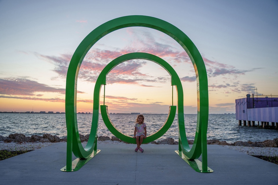 Green powder-coated steel loop sculpture with a child seated inside at sunset on a waterfront park boardwalk, by The Urban Conga
