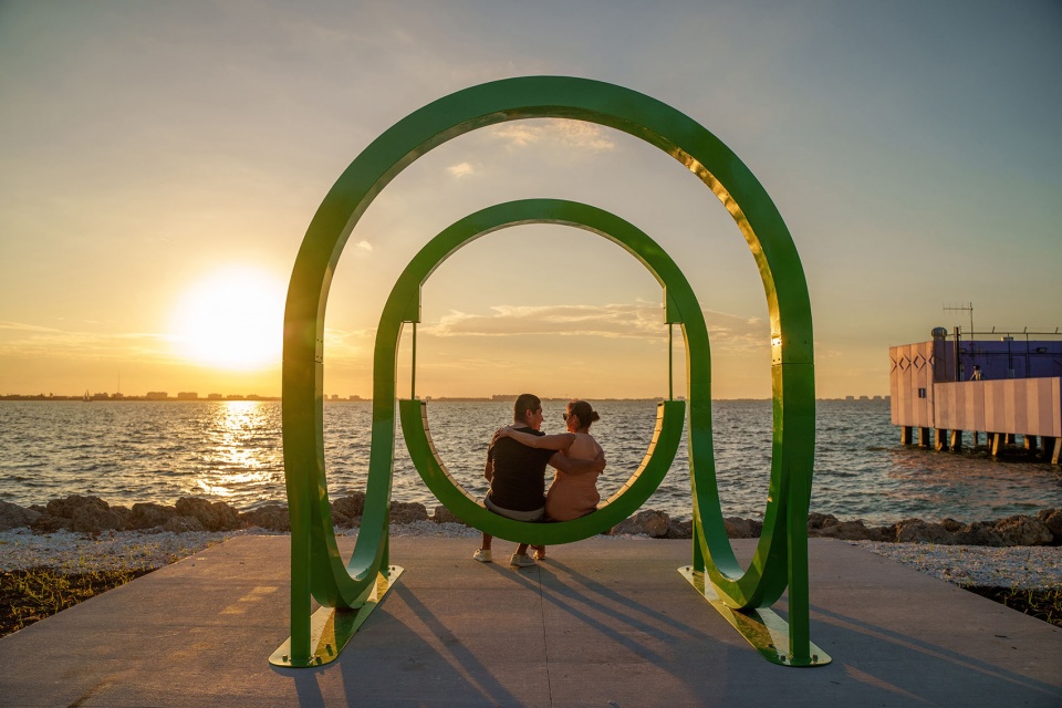 Two people sitting on Portal, a looped swing sculpture by The Urban Conga, with the sun setting inside the loop at The Bay Park, Sarasota