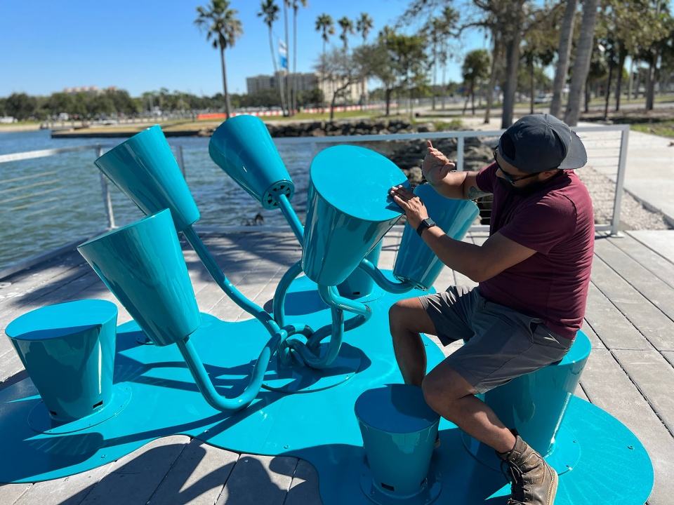 Visitor playing a blue powder-coated steel musical sculpture beside the water, playable art installation by The Urban Conga