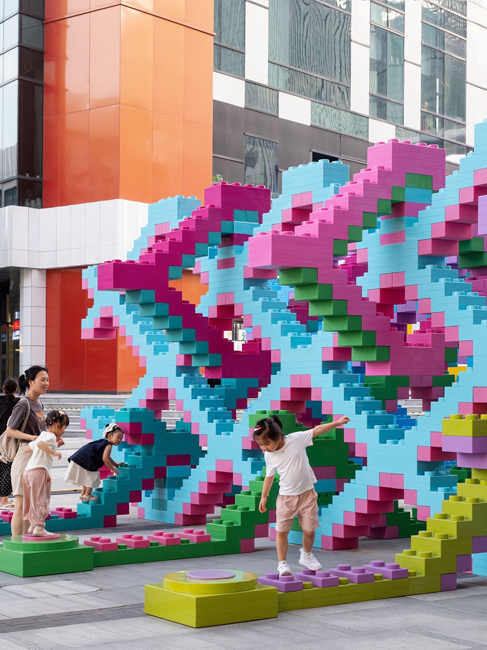 Oversized pastel toy brick sculpture with children climbing on stepping blocks in front of an office tower, Shanghai