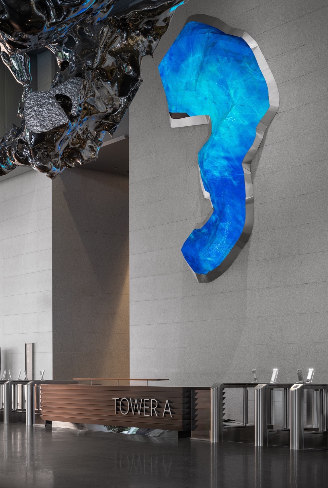 Detail of a hanging polished stainless steel sculpture above a lobby reception desk with a backlit blue metal-framed wall piece